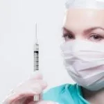 alt="Enfermera vestida de verde con mascarilla y paño en cabeza, sosteniendo una geringuilla mientras mira de frente"