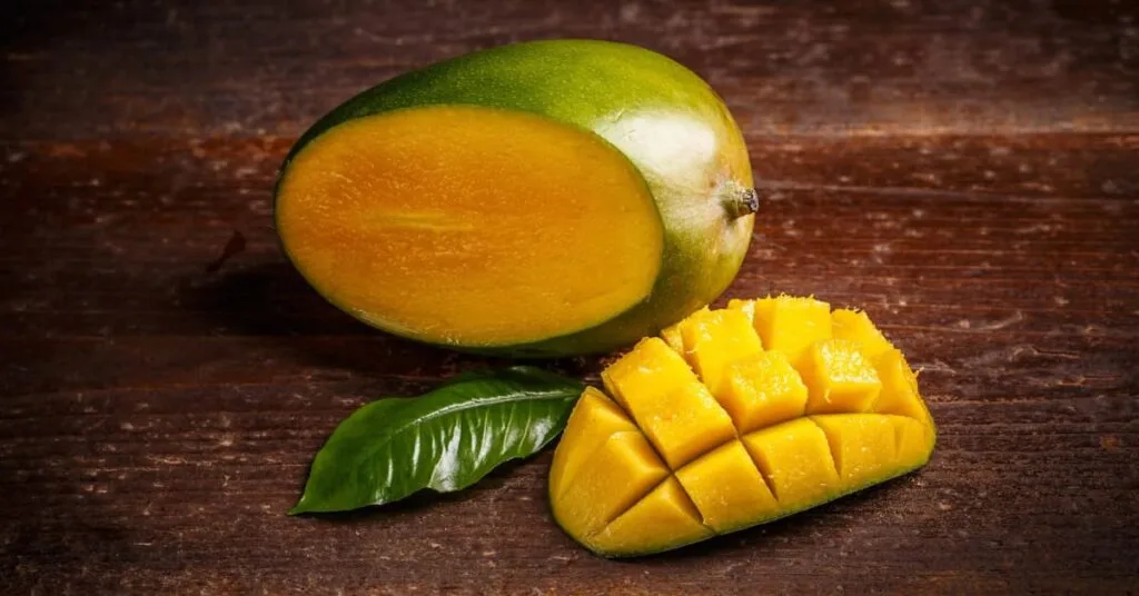 alt="Mango con la cascara verrdosa amrillenta con una rodaja cortada acentada al lado, y debajo se ve una hoja fresca"