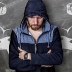 alt="Hombre recostado a la pared con los brazos cruzados, de fondo unos brazos pintados con crayon blanco simbolizando fortaleza"