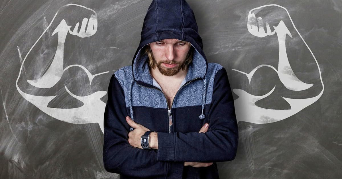 alt="Hombre recostado a la pared con los brazos cruzados, de fondo unos brazos pintados con crayon blanco simbolizando fortaleza"