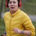 alt="Mujer asiatica con abrigo amarillo y audifonos rojos corriendo en medio del campo mientras mira diratamente a la camara"