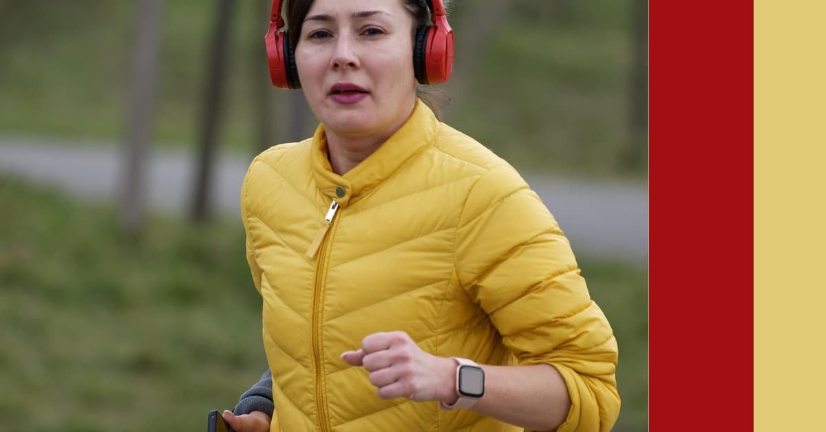 alt="Mujer asiatica con abrigo amarillo y audifonos rojos corriendo en medio del campo mientras mira diratamente a la camara"