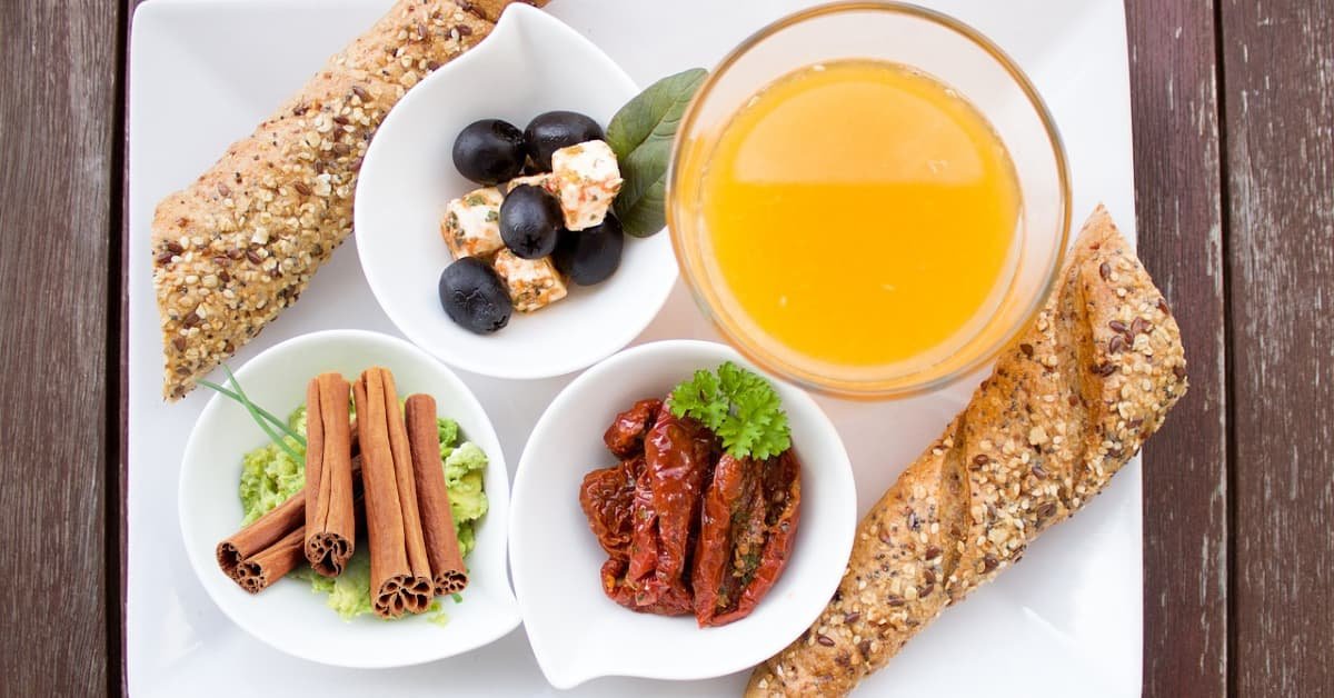 alt="Desayuno saludable en plato cuadrado blanco, frutas, yogures, jugos naturales y tostadas de pan"