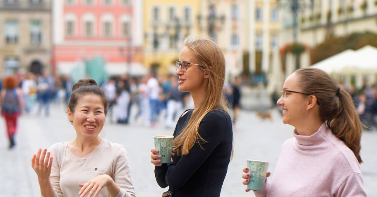 alt="Amigas en medio de una plaza llena de personas, relajadas tomando cafe y conversando"