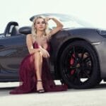 alt="Mujer agachada recostada a carro de lujo, vistiendo un vestido elegante rojo"