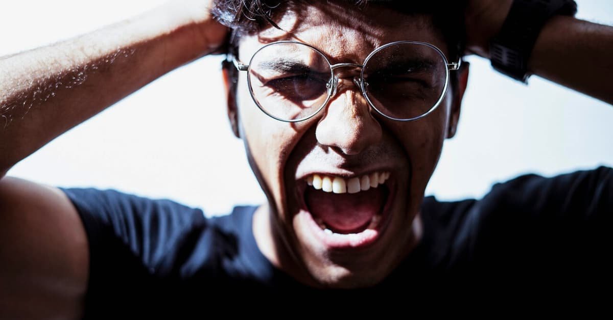 alt="Con las manos en la detras de la cabeza y griitando desesperado aprece en una habitacion vacia un hombre joven con una camiza verde,,lleva puesto unos Espejuelos de leer y un reloj"