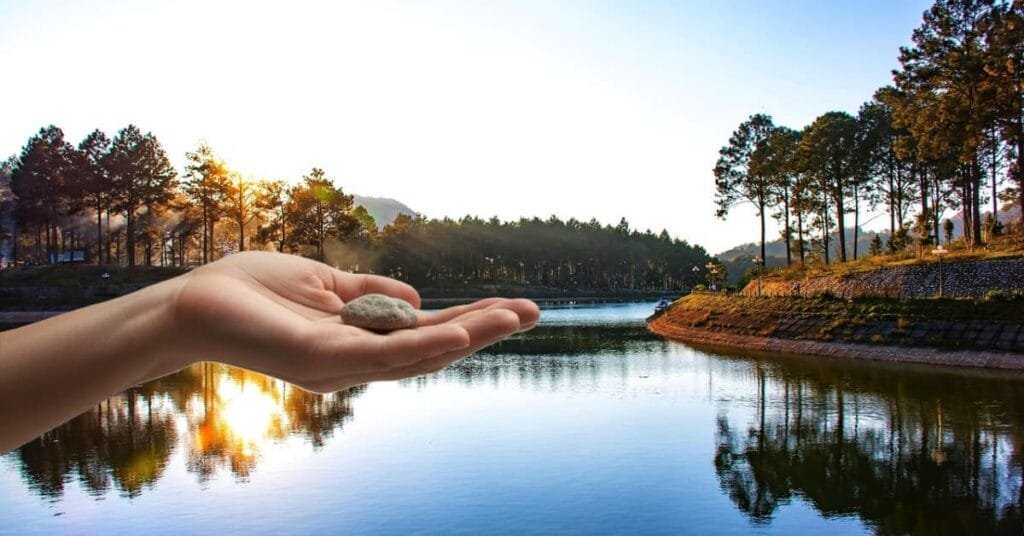 alt="una mano abierta extendida con una pierda en la plama. al fondo se ve un cielo con unos rallos de sol callendo sobre montañas Bosques y un Rio"