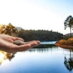 alt="una mano abierta extendida con una pierda en la plama. al fondo se ve un cielo con unos rallos de sol callendo sobre montañas Bosques y un Rio"