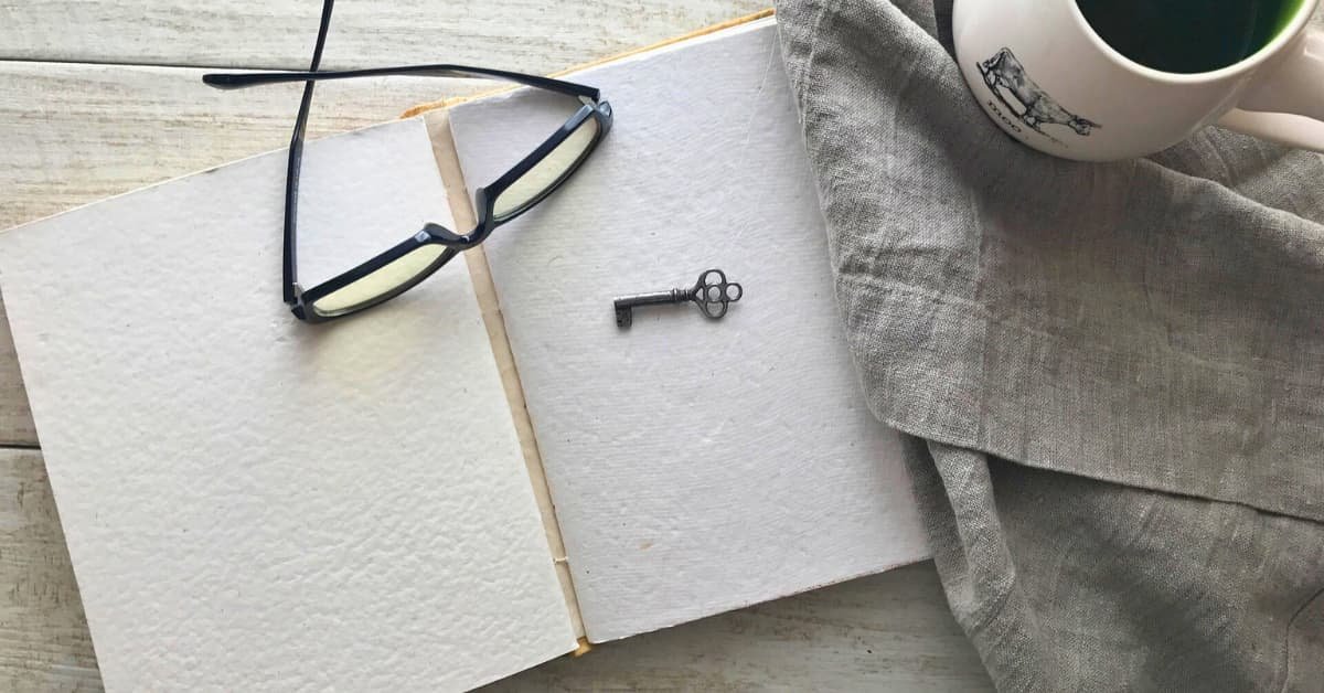 alt="sobre una mesa de tablas blancas estan colocados y organizados una libreta de apuntes abierta, con unas gafas de leer y una llave antigua encima. Al lado sobre un mantel de tela una taza blanca llena de cafe humeante."