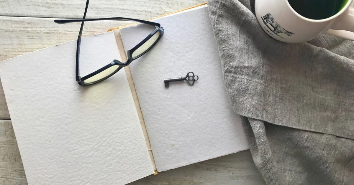 alt="sobre una mesa de tablas blancas estan colocados y organizados una libreta de apuntes abierta, con unas gafas de leer y una llave antigua encima. Al lado sobre un mantel de tela una taza blanca llena de cafe humeante."