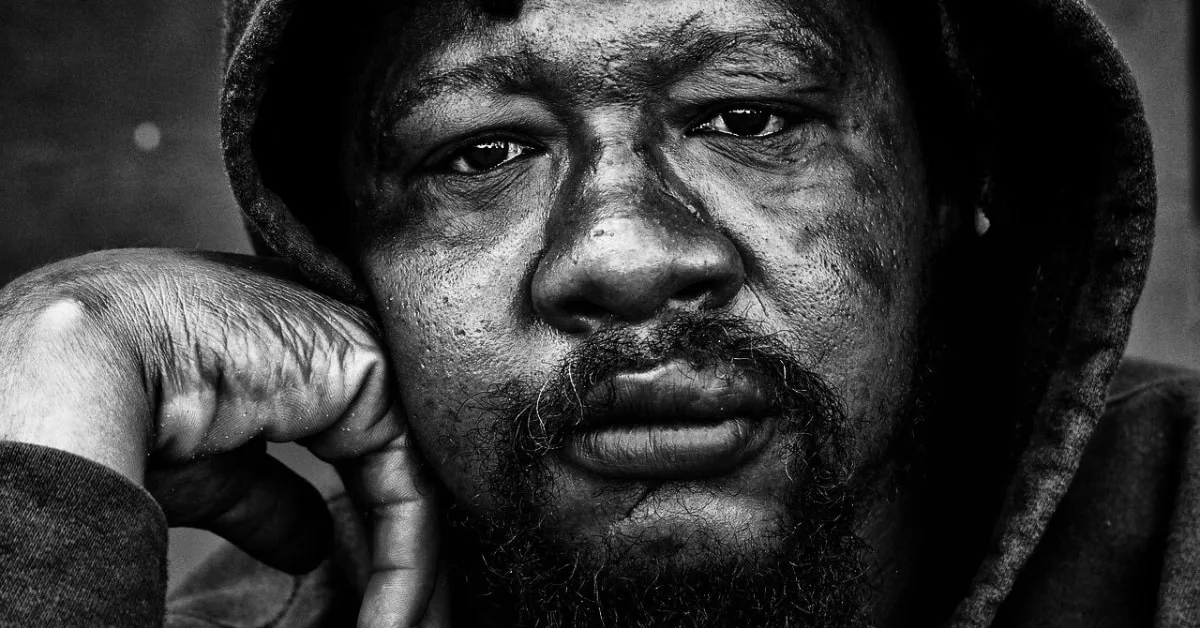 alt="Retrato en blanco y negro de un hombre con barba y capucha, apoyando la mejilla en su mano, con una mirada profunda y pensativa."