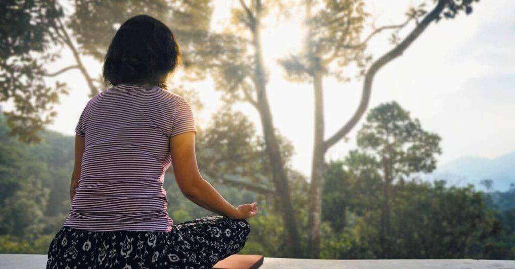 alt="mujer sentada de esplada, meditando a las orillas de un bosque intenso lleno de paz"