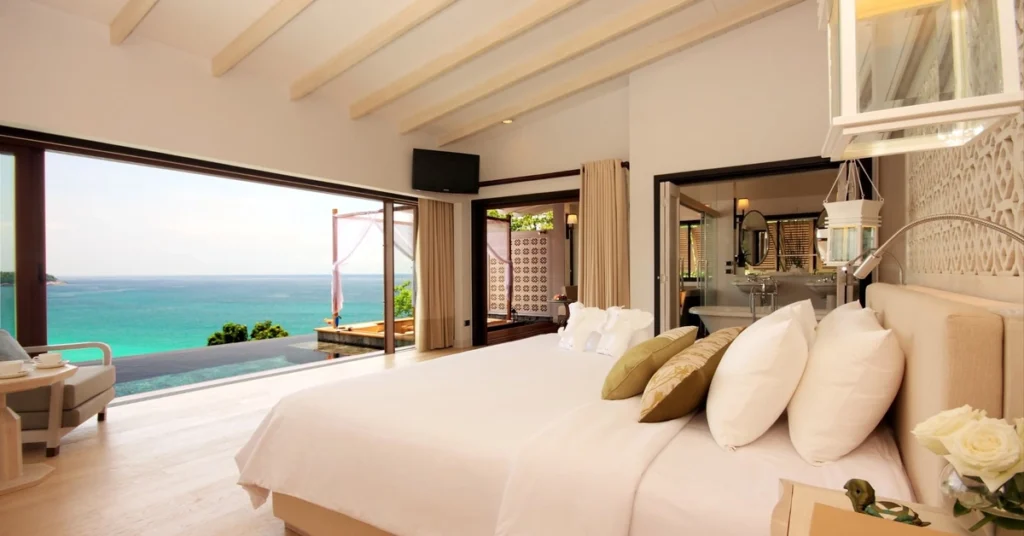 Alt="Habitacion lujosa en la playa, espaciosa minimalista con cama enorme, y un ventabal que se ve el mar color verde turqueza atravez"