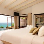 Alt="Habitacion lujosa en la playa, espaciosa minimalista con cama enorme, y un ventabal que se ve el mar color verde turqueza atravez"