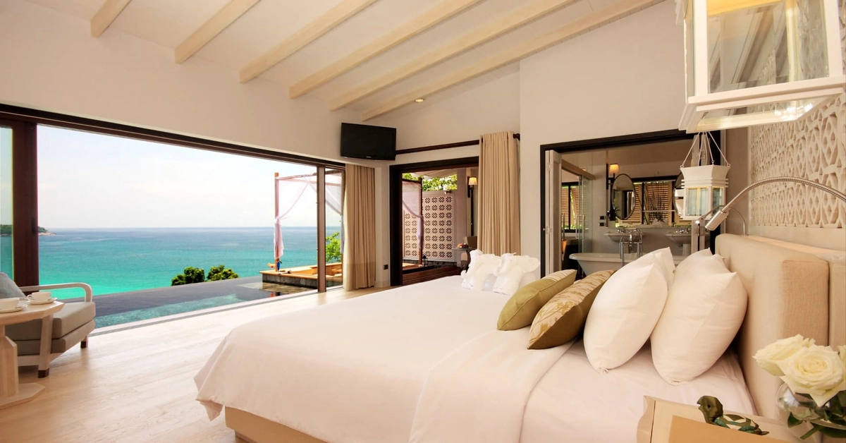 Alt="Habitacion lujosa en la playa, espaciosa minimalista con cama enorme, y un ventabal que se ve el mar color verde turqueza atravez"