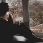 alt="Silueta de una mujer descalza sentada en el alféizar de una ventana interior, sosteniendo una taza blanca y mirando hacia afuera, a un paisaje invernal con árboles cubiertos de nieve."